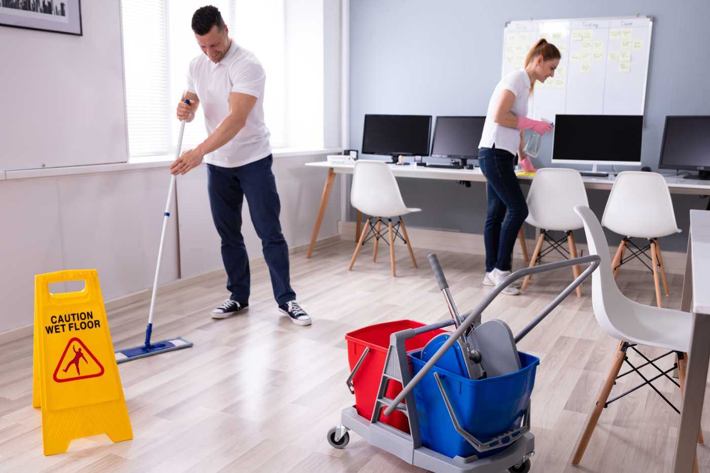 Two Janitors Cleaning A Desk And Mopping The Floor Of An Office